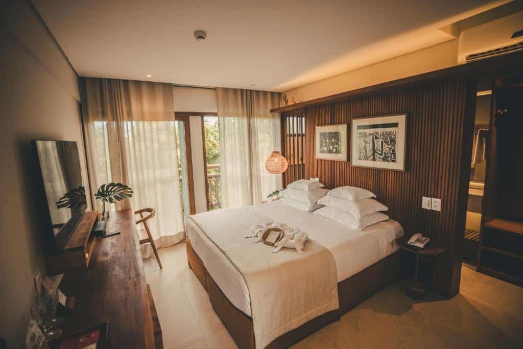 Quarto do Refúgio da Vila. Uma cama de casal com seis travesseiros do lado direito. De frente uma raque com televisão, mesa de trabalho e uma cadeira. No fundo, a sacada do quarto.