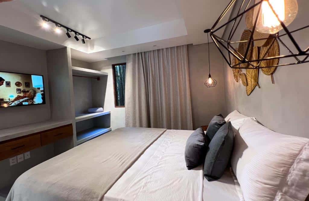 Quarto do Refugio Do Morro. Uma cama de casal do lado direito com um abajur de cada lado. Do lado esquerdo uma televisão e prateleiras. No fundo a janela com cortina. Foto para ilustrar post sobre airbnb em Morro de São Paulo.