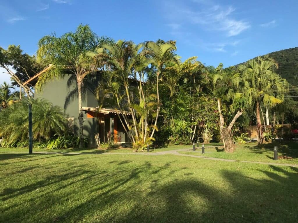 Área externa do Residencial As Corujas. Uma casa está atrás de várias árvores e tem um grande gramado como quintal.