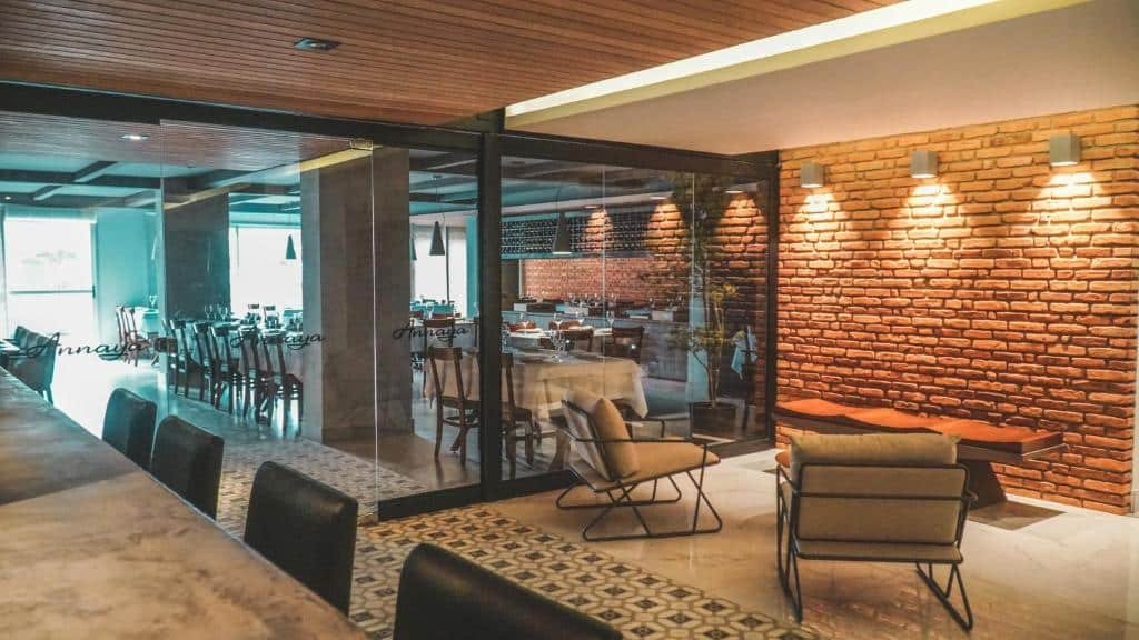 Entrada do restaurante do hotel Annaya Suítes. Na parte de fora do restaurante há um balcão no lado esquerdo com cadeira e duas poltronas no lado direito. É possível ver o restaurante através da parede de vidro. Lá dentro estão dispostas diversas mesas com cadeiras.