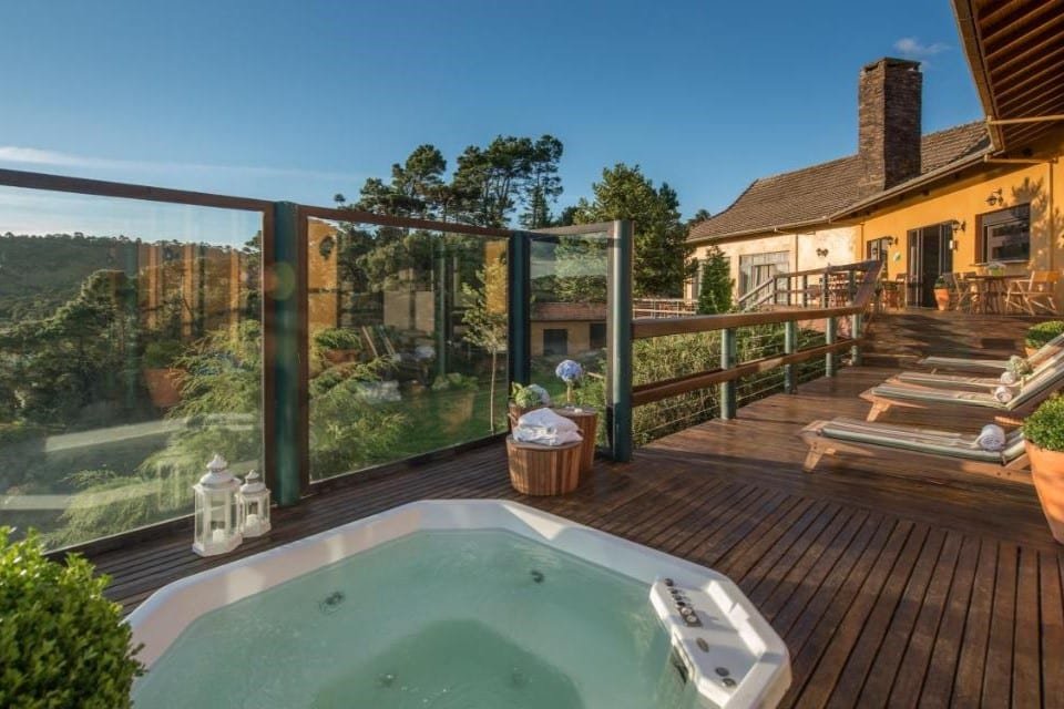 Um deck de madeira com hidro, espreguiçadeiras, vasos de plantas e cercado por grades de vidro oferece vista para uma paisagem florestal no Rio do Rastro Eco Resort.