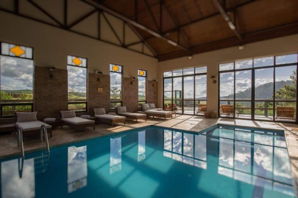 Área de piscina coberta com espreguiçadeiras ao longo das janelas e portas de vidro proporcionando vista para as montanhas e o céu. Esse é o Rio do Rastro Eco Resort, um dos hotéis fazenda em Santa Catarina.