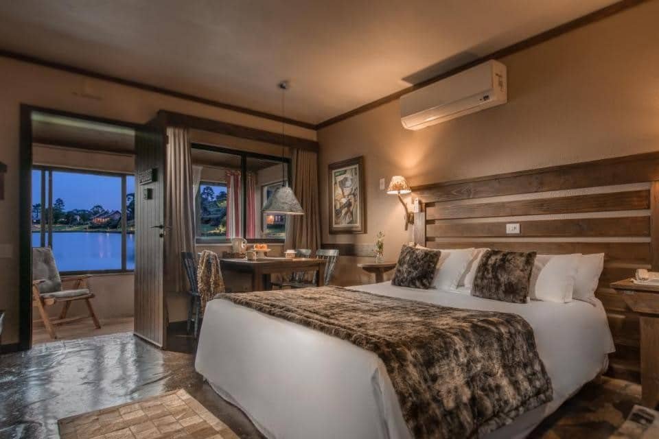 Um quarto aconchegante do Rio do Rastro Eco Resort com uma cama grande, cobertores de pele, uma pequena mesa de jantar e uma janela com vista para um lago ao anoitecer.