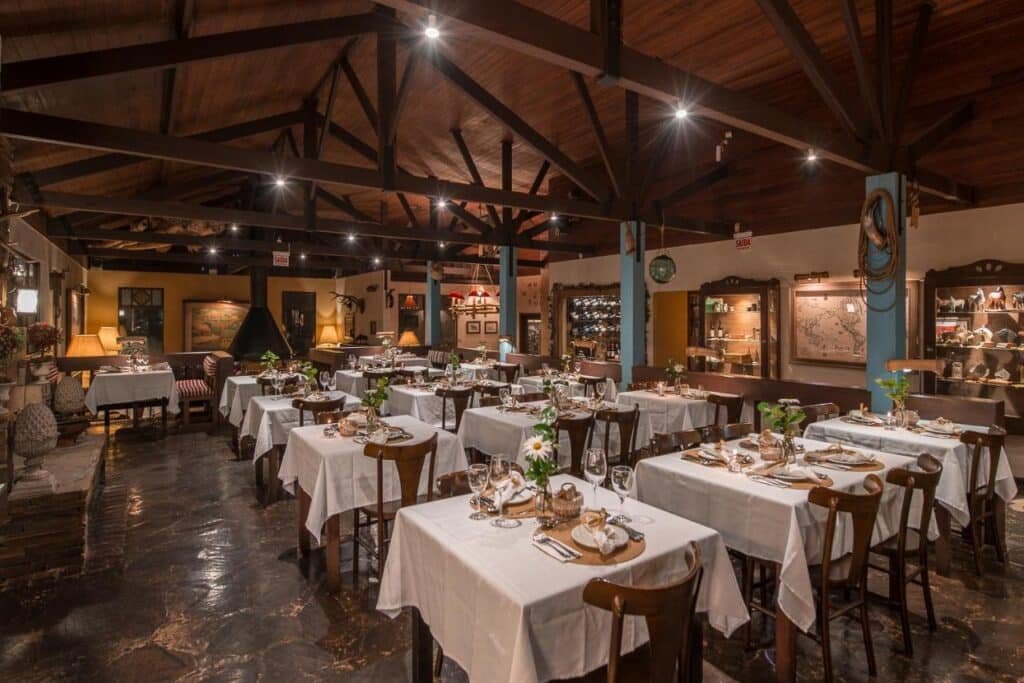 Um restaurante aconchegante com vigas de madeira, mesas com toalhas brancas e iluminação suave, criando uma atmosfera acolhedora. Esse é o Rio do Rastro Eco Resort, um dos hotéis fazenda em Santa Catarina.