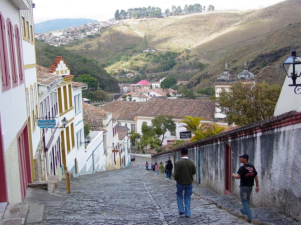 Uma rua íngreme de paralelepípedos em uma cidade montanhosa com prédios coloniais coloridos. Pessoas estão andando pela rua, e colinas estão ao fundo.