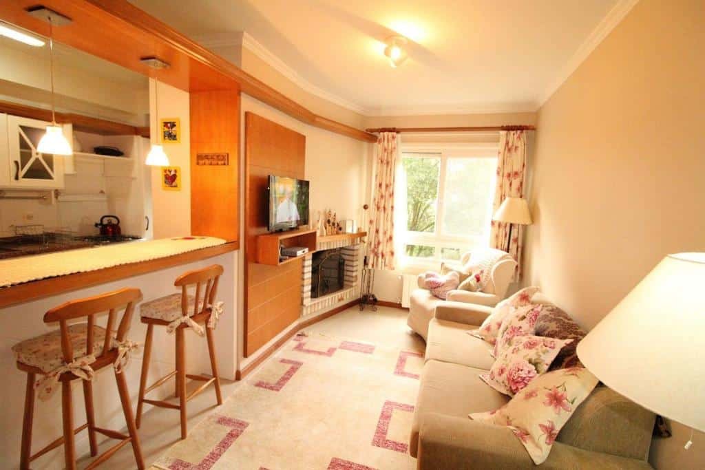 Sala de estar na Araucárias 302, uma sala com um sofá e uma poltrona ao lado de uma sacada com vista para as árvores, dois abajures, uma lareira, uma televisão e um balcão com duas cadeiras