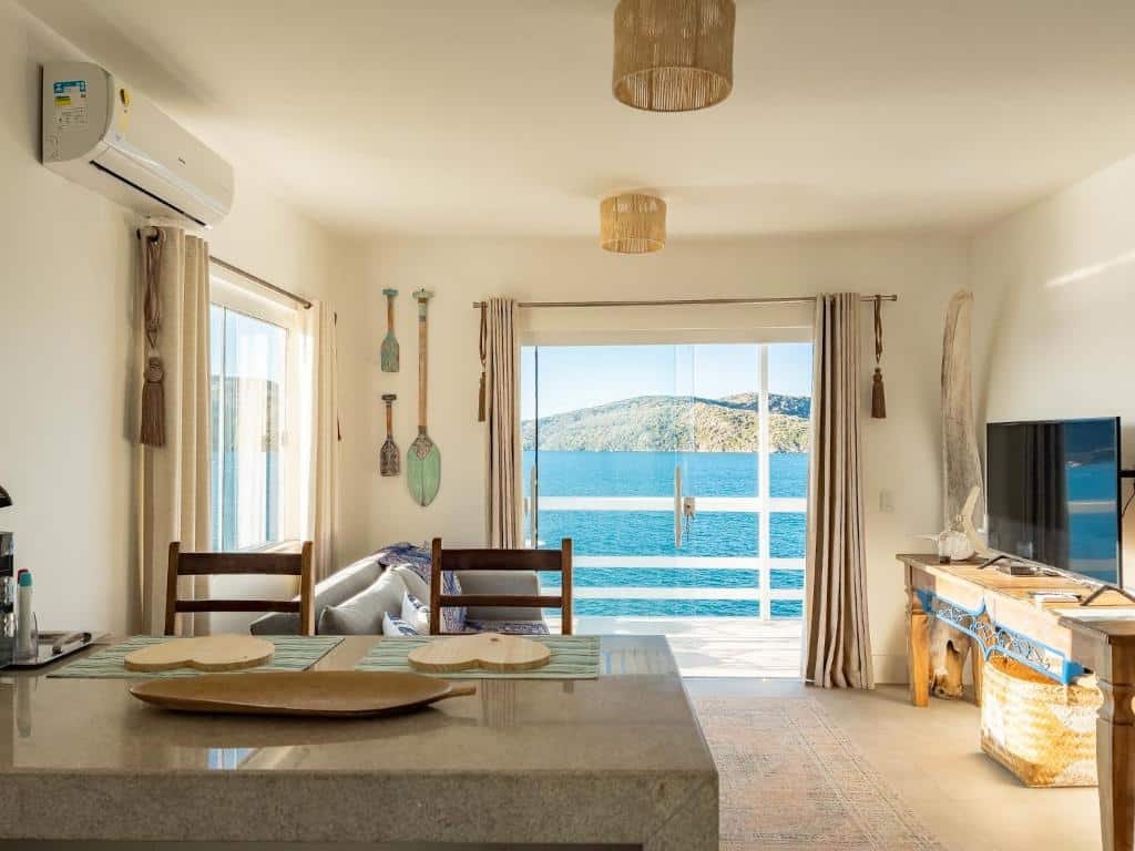 Sala de estar do airbnb Casa Santorini Terrace. No lado direito da sala há um rack com uma TV em cima, em frente ao rack, no lado esquerdo, há um sofá e uma janela de vidro. No fundo há uma porta e vidro grande que dá acesso a varanda e vista para o mar. Já na frente da imagem há uma mesa de granito com duas cadeiras.