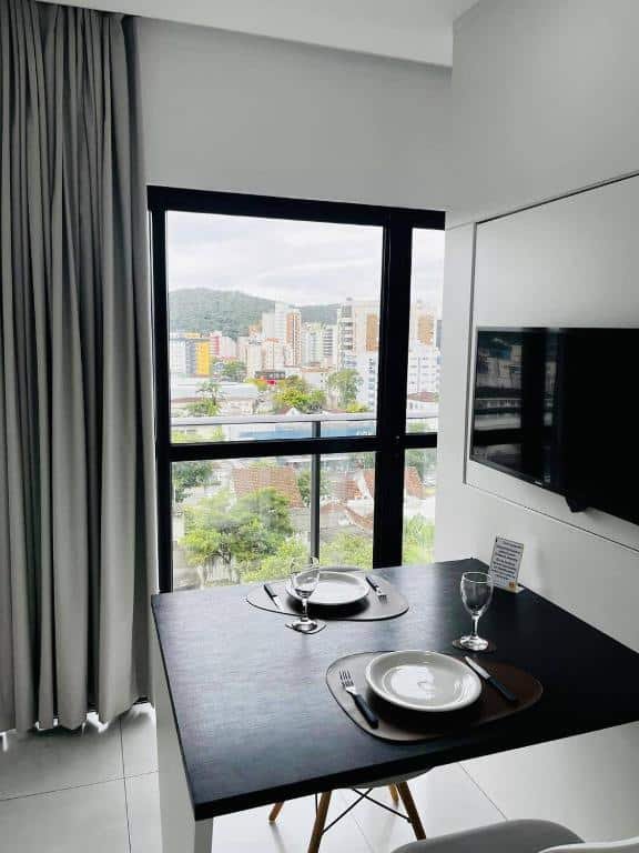 Sala de jantar e de estar do MaxLoft mostrando uma mesa com dois lugares, equipada com dois pratos e duas taças em cima. Ao fundo da imagem é possível ver a sacada particular do apartamento com vista para a cidade.