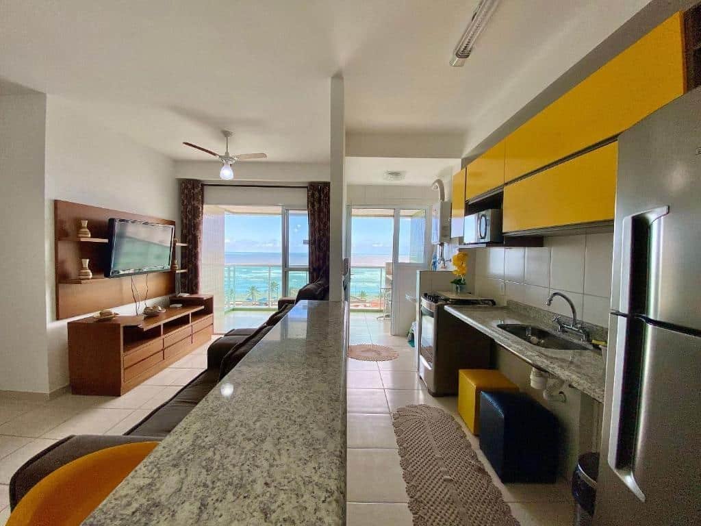 Apartamento com sala com sofá, móvel de madeira com tv no lado esquerdo, bancada no meio e no lado direito cozinha com geladeira, pia, fogão e armários. No fundo dos dois cômodos há uma enorme varanda com vista para o mar.