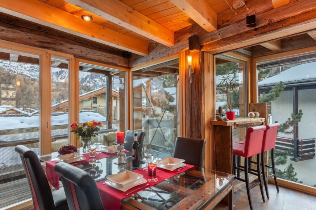 Sala de refeição do Chalet Schwalbennest com uma mesa de vidro quadrada com quadro cadeiras do lado esquerdo a frente as parede da sala são de vidro que oferece vista para o jardim coberto de neve.