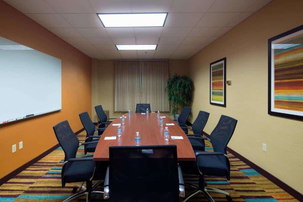 Imagem de uma sala de reuniões no Fairfield Inn & Suites. A sala é espaçosa e bem iluminada, com uma grande mesa retangular no centro, cercada por cadeiras confortáveis. Na parede oposta à mesa, há um quadro branco e uma tela de projeção.