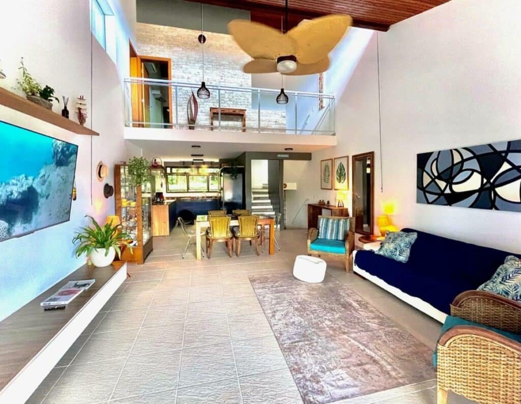 Sala de estar do Delícia de casa em Barra do Una a 5 min de Juquehy! a frente com sofá do lado direito da imagem em frente ao sofá uma cômoda com TV presa na parede. Ao fundo da sala uma mesa com quatro cadeiras.