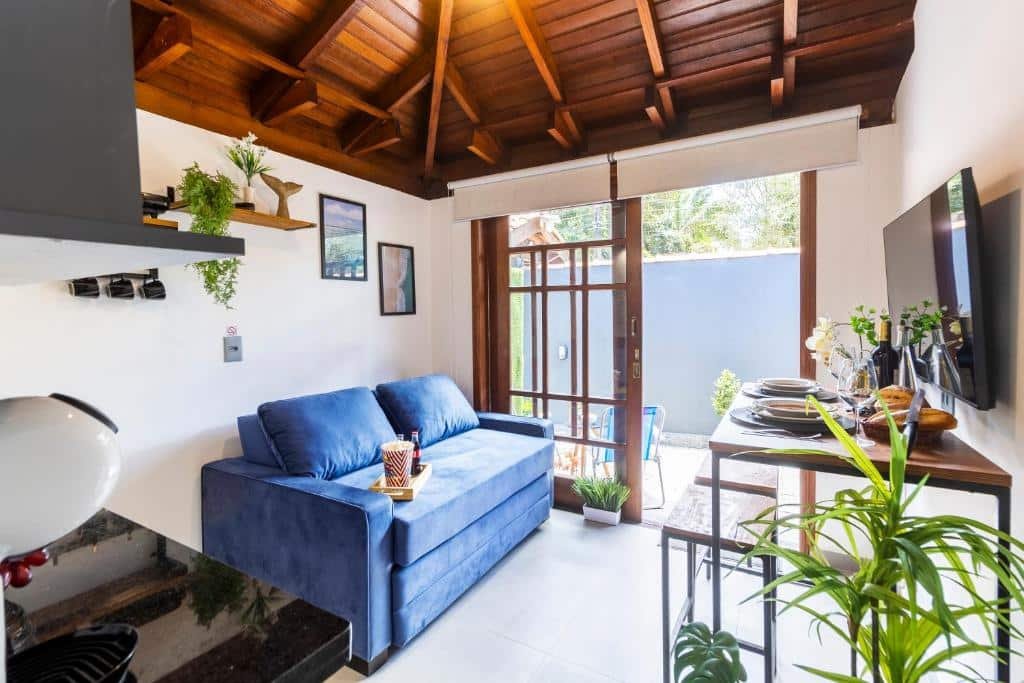 Sala de estar do Flats Una’s Corner – Novos – Ar Condicionado com sofá do lado esquerdo da imagem e em frente ao sofá uma mesa com dois bancos com uma TV presa na parede.
