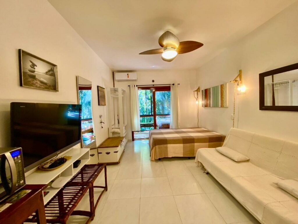 Sala do Housing 31 – Cond. Mata Azul com sofá a frente do lado direito da imagem em frente ao sofá uma cômoda com TV e do lado esquerdo do sofá uma cama.