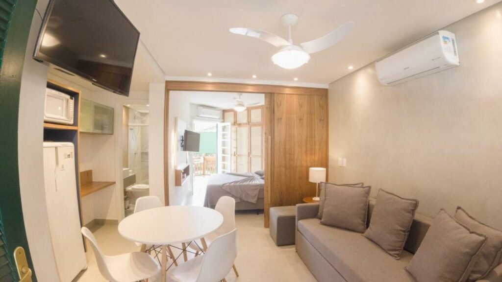 Imagem do ambiente do Porto Paúba Flat 116 com sala de estar a frente com sofá do lado direito da imagem, em frente ao sofá uma mesa redonda com quatro cadeiras e do lado direito da mesa uma cozinha compacta. Representa airbnb na praia de Santiago.