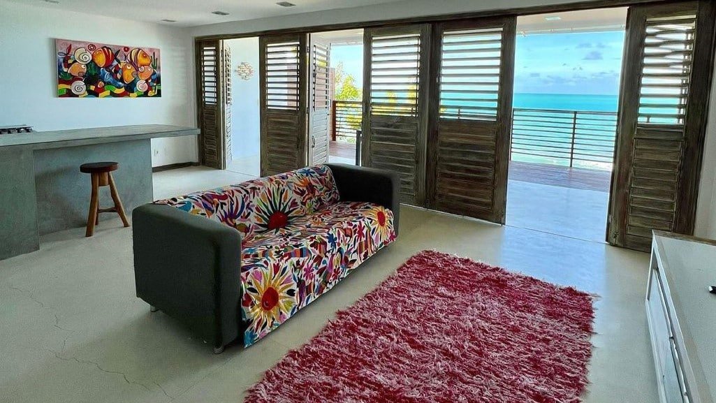 Sala de estar da Pousada ResDelMar. Um balcão e um banquinho está no fundo, um sofrá está no meio e logo em frente a ele há um tapete felpudo. Em frente ao tapete há um rack. Na parede ao lado de tudo isso estão várias portas levando até a varanda, que fica de frente para o mar.