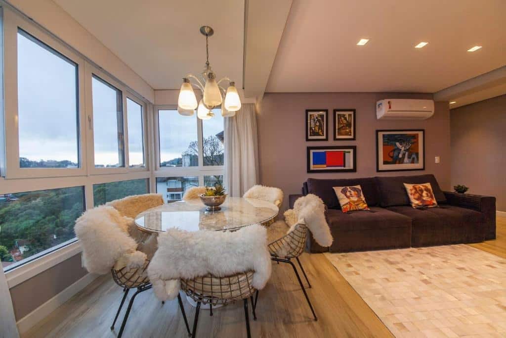 Sala de estar do Residencial Via Florida Gramado, com uma janela ampla que oferece vista para as árvores, uma mesa redonda com seis lugares, um lustre, um sofá, um tapete e um ar-condicionado na mesma parede que há três quadros