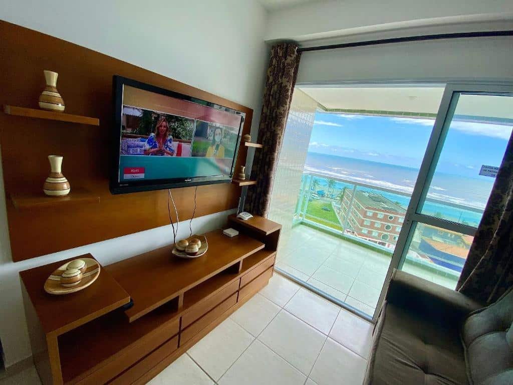 Sala de apartamento com móvel de madeira com televisão no lado esquerdo, sofá no lado direito e varanda com vista para o mar ao fundo.