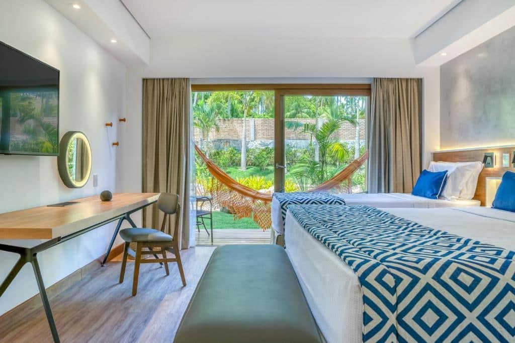 Quarto de um apartamento do Salinas Maragogi, de 32 m², com duas camas e, à esquerda, tem um TV suspensa e uma mesa com uma cadeira. Há uma varanda, separada por uma porta de vidro, contendo nela uma rede e vista para um jardim