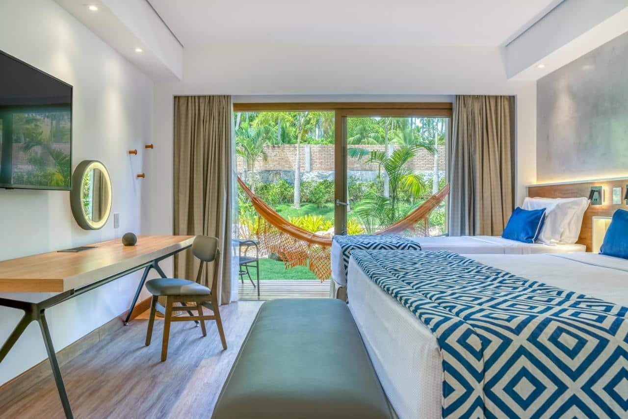 A imagem mostra um quarto do Salinas Resort, em Maragogi, bem iluminado e moderno. No centro, há uma cama de casal e ao lado outra de solteiro, ambas com uma colcha branca e uma coberta decorativa com padrões azuis e brancos acima. Uma rede laranja está pendurada na frente de uma grande porta de vidro que leva a uma varanda externa com vista para um jardim tropical. À esquerda da cama, há uma mesa de trabalho de madeira com uma cadeira moderna, um espelho redondo e uma TV na parede acima. À direita, um painel de madeira serve de cabeceira. O chão é de madeira clara, e a decoração geral é clean e convidativa, com um toque de cor vibrante fornecido pela rede.