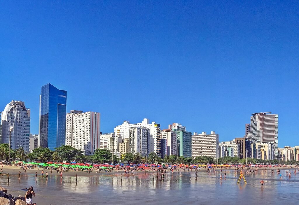 Uma praia bem extensa com muitos guarda-sóis coloridos, pessoas andando e, logo atrás da praia, há muitos prédios comerciais