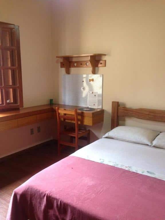 Quarto do Sefiroh Hostel. No lado direito há uma cama de casal e no lado esquerdo há uma mesa em formato de L com uma cadeira, uma prateleira em cima e uma janela no lado esquerdo. Imagem para ilustrar o post hotéis em Viçosa.