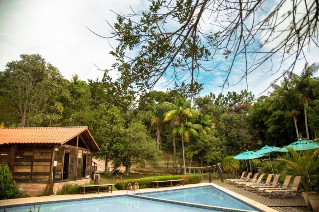Uma área de piscina tranquila com espreguiçadeiras, guarda-sóis, árvores e uma casa de madeira em um ambiente florestal exuberante. Esse é o Serra do Ibitipoca Hotel de Lazer, um dos hotéis fazenda em Minas Gerais.