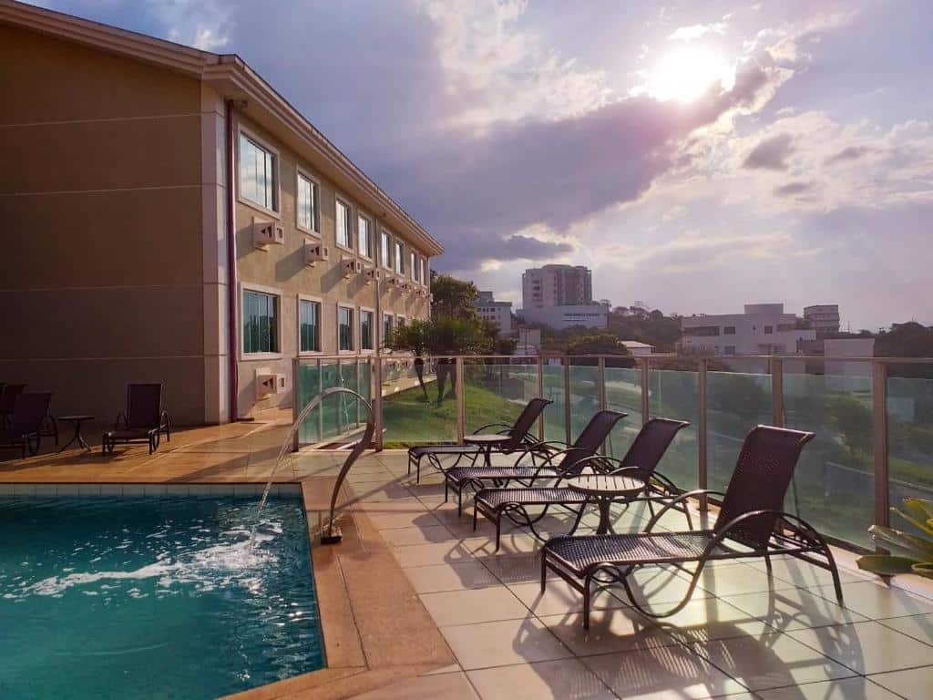 área da piscina do Serra Palace Hotel em Ouro Branco, perto de Congonhas, com quatro espreguiçadeiras beirando em frente a piscina, uma cascata de metal em frente às cadeiras com caimento abundante de água na piscina. e um amplo guarda-corpo de vidro separado a área da piscina da rua mais abaixo. Ao fundo é possível enxergar o hotel com várias janelas e ar-condicionados