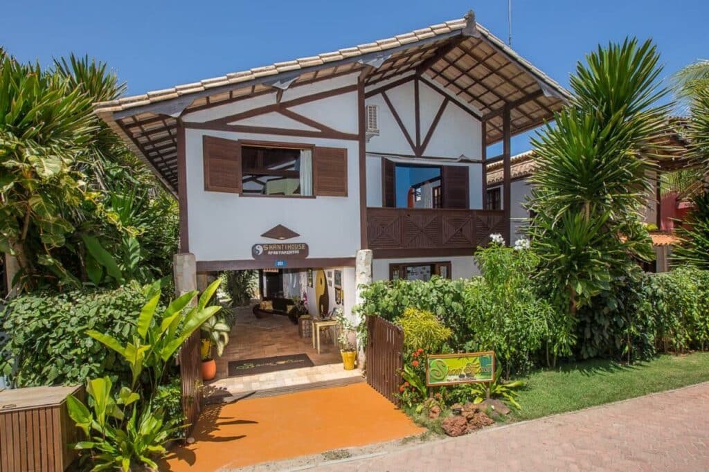 Vista de fora da casa de temporada Shanti House Apart. Ela possui dois andares, é toda pintada de branca mas possui janelas e telhado marrons. Ela é toda rodeada por árvores e plantas.