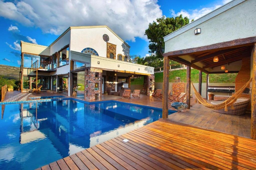 área da piscina do Sítio Jatobá com uma churrasqueira coberta à direta, uma área para rede, várias cadeiras espreguiçadeiras de madeira e a construção em pedra da pousada ao fundo, pintada de branco.