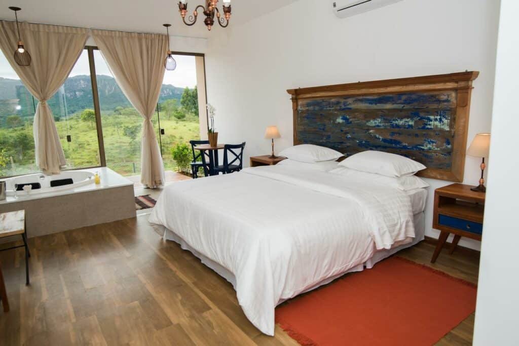 quarto espaçoso da pousada Solar da Serra Tiradentes com uma cama de casal grande, uma mesa redonda pequena de madeira com duas cadeiras pretas, e uma banheira de hidromassagem redonda com espaço para duas pessoas. Ao fundo do quarto é possível apreciar a vista dos vales numa parede inteira de vidro transparente.