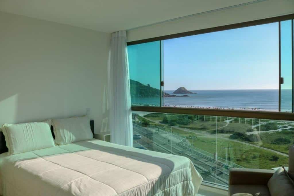 Quarto do Sophia II Residence Inn. No lado esquerdo há uma cama de casal encostada na parede, em frente a cama é possível ver uma parte de uma poltrona e no lado direito a parede do quarto é toda feita de vidro, a parte superior está aberta e oferece vista para o mar.