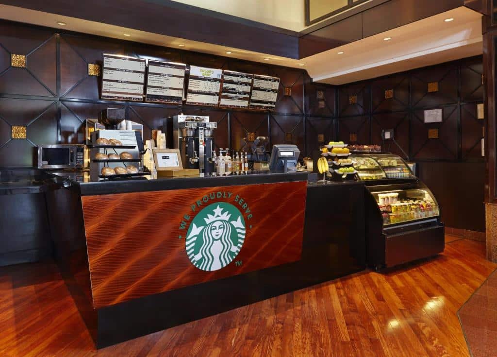 Imagem de uma unidade do Starbucks localizada no Hilton Newark Airport. O estabelecimento possui um design moderno e acolhedor. No balcão central, baristas estão prontos para atender os clientes. A vitrine exibe uma variedade de bebidas e alimentos disponíveis. Mesas e cadeiras estão dispostas ao redor do espaço para os visitantes desfrutarem de suas bebidas e lanches.