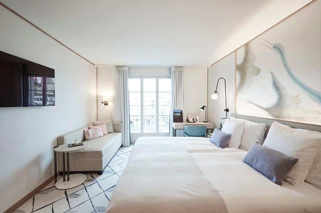 Quarto do Storchen Zürich Boutique Hotel, com decoração clean mesclando cores claras no tom branco, com uma cama grande, sofá, TV e mesa de trabalho com cadeira azul