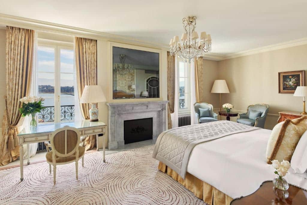 Foto de um quarto do Beau Rivage Geneve, para ilustrar o post de hotéis em Genebra. O quarto é bem iluminado, as paredes possuem coloração bege e o chão tem um carpete branco. A cama está de costas para a câmera da foto, no lado direito. E no canto inferior direito, no lado dela, percebe-se uma mesa de cabeceira marrom com um vaso de flores em cima. No canto direito do quarto, no fundo da foto, há duas poltronas azuis e uma mesa de centro entre elas, e um abajur de chão atrás. Na frente da cama, há uma lareira. E do lado esquerdo da lareira está uma mesa de escrivaninha branca com abajur e cadeira. Na frente, uma janela. O teto possui um lustre branco.