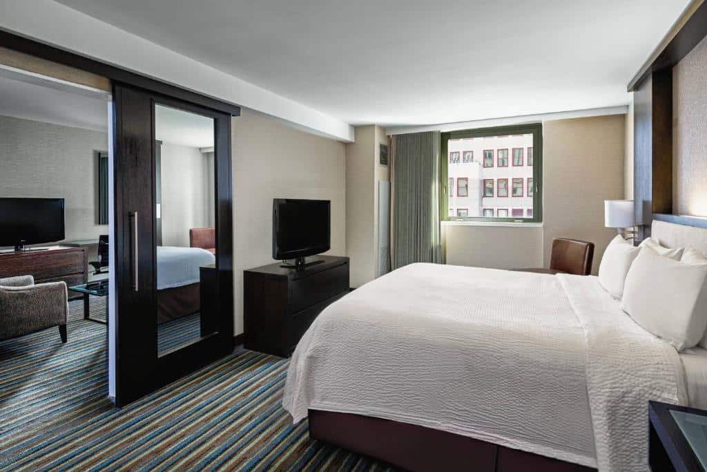 quarto com dois cômodos do Courtyard New York Manhattan/Times Square onde há uma cama de casal no lado direito da imagem, enquanto no lado esquerdo, dividido por uma parede e uma porta espelhada, está uma pequena sala de estar.