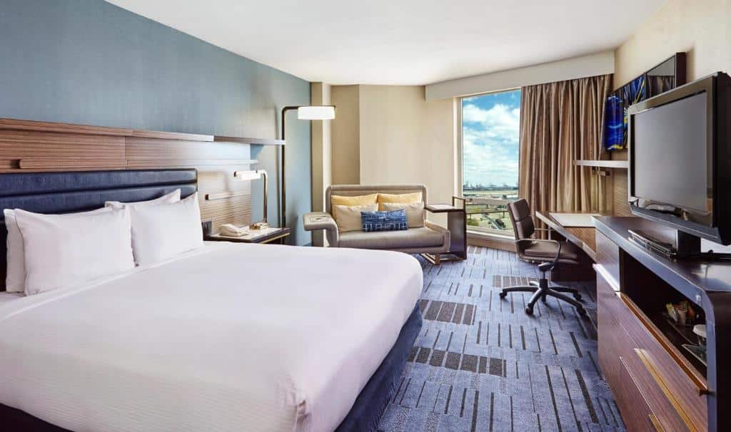 Imagem de uma suíte espaçosa no Hilton Newark Airport. A suíte apresenta uma decoração elegante com mobiliário luxuoso. Na área de estar, há um sofá posicionado à esquerda da imagem, ao lado de uma mesa de centro. Do lado direito, há poltronas confortáveis. Na área do quarto, há uma cama king-size posicionada no centro. O ambiente é moderno e acolhedor, oferecendo conforto e conveniência aos hóspedes. O Hilton Newark Airport é um hotel próximo ao aeroporto de Newark.