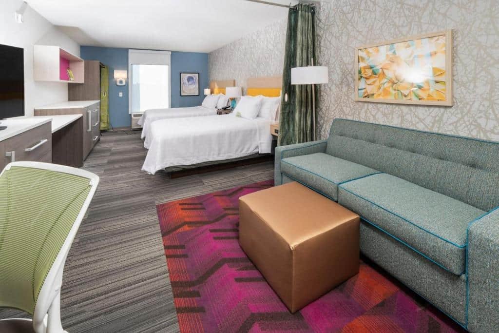 Imagem de uma suíte no Home2 Suites Newark Airport. A suíte é espaçosa e bem iluminada, com uma área de estar separada por uma parede parcial. Na área do quarto, há uma cama king-size com lençóis brancos e travesseiros macios. À esquerda da cama, há uma mesa de cabeceira com uma luminária de leitura. Na área de estar, há um sofá e uma mesa de centro. Próximo à janela, encontra-se uma escrivaninha com uma cadeira. O Home2 Suites Newark Airport é um dos hotéis perto do aeroporto de Newark.