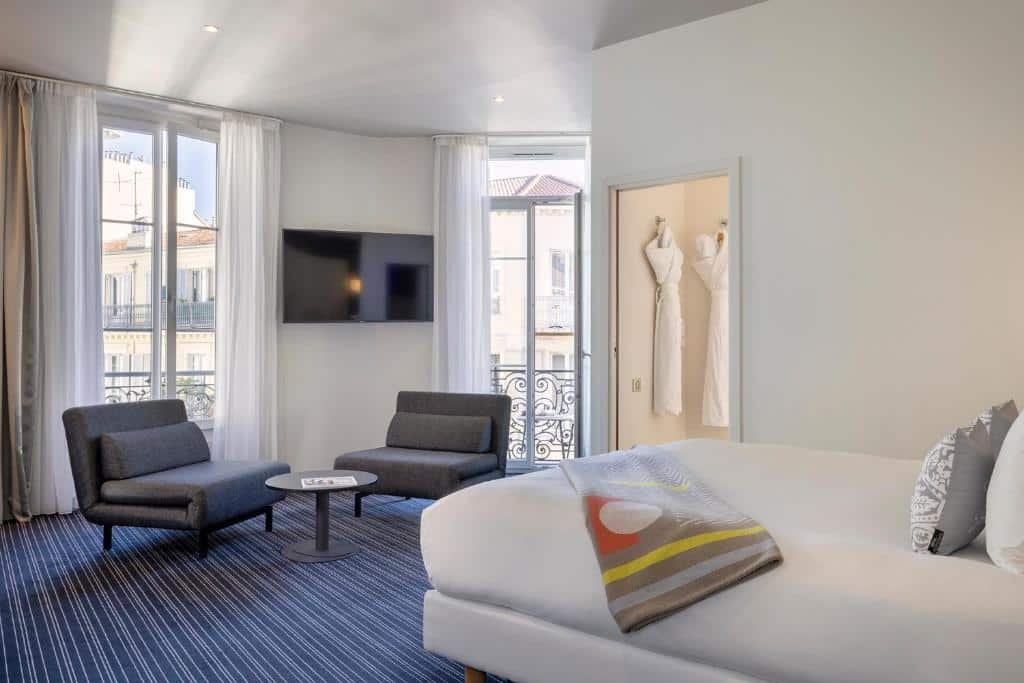 Quarto do Hotel 66 Nice. A cama está encostada na parede direita do quarto, ao lado direito da cama está o banheiro e na frente há duas poltronas, uma mesa de centro, uma TV apoiada na parede e duas portas de vidro com cortina que dão para a varanda. Imagem para ilustrar o post hotéis em Nice.