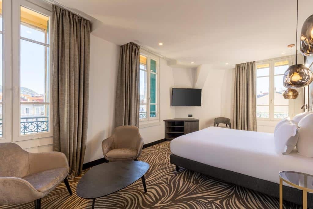 Quarto do Hôtel Apollinaire Nice. A cama está encostada na parede do lado direito do quarto, na parede dos fundos há uma janela grande, uma cadeira, uma cômoda e uma TV apoiada na parede. No lado esquerdo do quarto há outras duas janelas grandes, duas poltronas e uma mesa de centro. Imagem para ilustrar o post hotéis em Nice.
