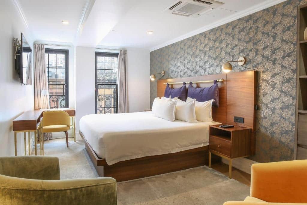 suíte do Hotel Scherman com uma cama de casal no lado direito, com uma parede totalmente coberta por papel de parede estampado. Ao fundo há duas janelas altas com acesso a varanda privativa. No canto esquerdo superior é possível ver uma mesa de madeira de canto com uma cadeira amarela estofada