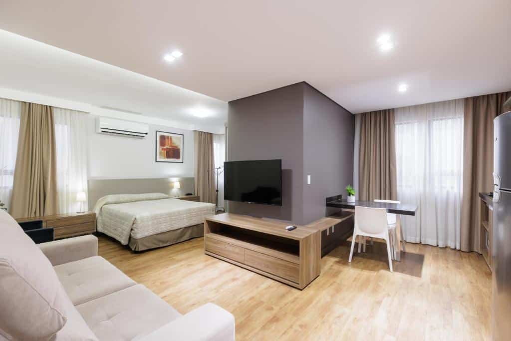 quarto no Le Village Flats Hotel e Business com uma cama de casal à esquerda, um sofá cama no canto inferior esquerdo da imagem, uma televisão de plasma em frente ao sofá e uma área de cozinha, com mesa para dois lugares e uma geladeira ao lado, no lado direito da imagem.