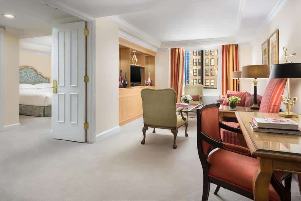 apartamento do Michelangelo Hotel com uma sala de estar à direita, onde há uma sofá e duas poltronas clássicas dispostas uma de frente para outra, com uma pequena mesa de centro entre elas. No lado esquerdo é possível ver uma porta aberta que dá acesso ao quarto, onde há uma cama de casal.