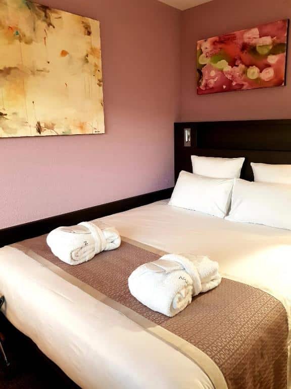 Foto de um quarto no Nash Pratik Hotel, para ilustrar o post de hotéis em Genebra. Há uma cama de casal, que ocupa maior espaço da foto. Há dois roupões em cima. As paredes são rosa e possuem dois quadros enfeitando.