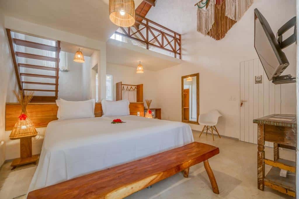 Suíte da Pousada Rota Ecológica, de 34 m², com cama de casal com uma flor vermelha em cima, 2 abajures artesanais (um de cada lado da cama) acesos, TV e escada dando acesso a uma área superior com rede