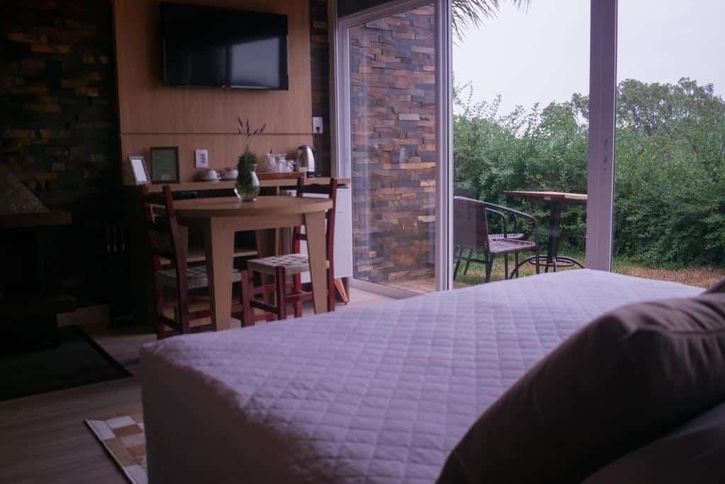 suíte da Pousada Cantelli, com cama de casal, mesinha com duas cadeiras, rack com chaleira e xícaras, além de TV presa à parede