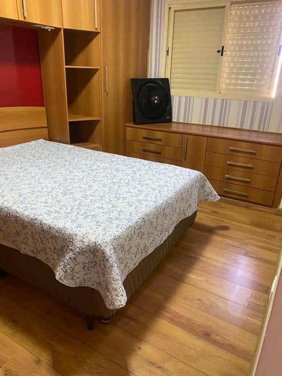 Quarto com chão e armários de madeira, janela no lado direito, ventilador em cima de móvel de madeira embaixo da janela e cama de casal centralizada. Imagem para ilustrar o post pousadas em Mongaguá.