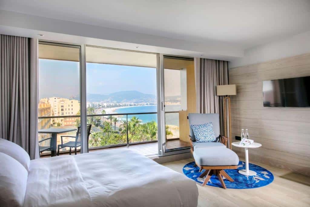 Quarto do Radisson Blu Hotel Nice. A cama está encostada na parede do lado esquerdo do quarto, em frente há uma poltrona, uma mesinha de centro redonda, um abajour grande de chão e a TV está presa a parede. A parede ao lado da cama é toda de vidro e dá acesso a varanda que contém uma mesa, duas cadeira e tem vista para a praia. Imagem para ilustrar o post hotéis em Nice.