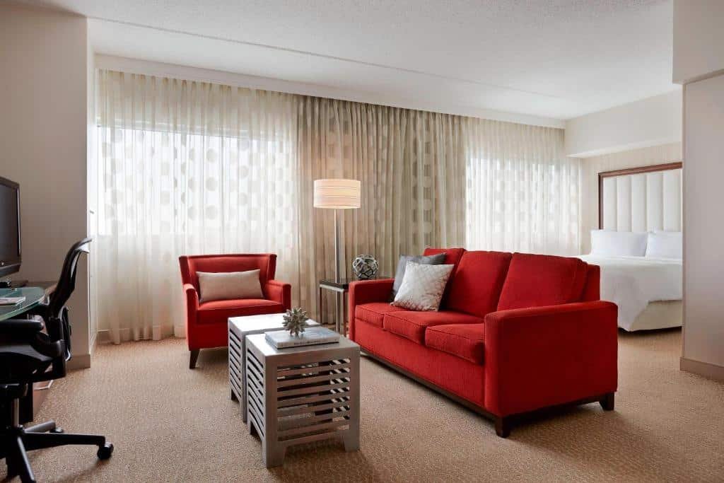 Imagem de uma suíte espaçosa no Renaissance Newark Airport Hotel. A suíte apresenta uma decoração sofisticada e elegante, com móveis de alta qualidade. À esquerda, encontra-se uma área de estar com um sofá confortável e uma mesa de centro. Ao centro, há uma cama king-size com lençóis impecáveis e travesseiros macios. À direita, há uma escrivaninha bem iluminada com uma cadeira ergonômica para trabalho ou estudo. O ambiente é luxuoso e convidativo, proporcionando uma estadia confortável aos hóspedes. O Renaissance Newark Airport Hotel é um hotel próximo ao aeroporto de Newark, oferecendo elegância e comodidade aos visitantes.