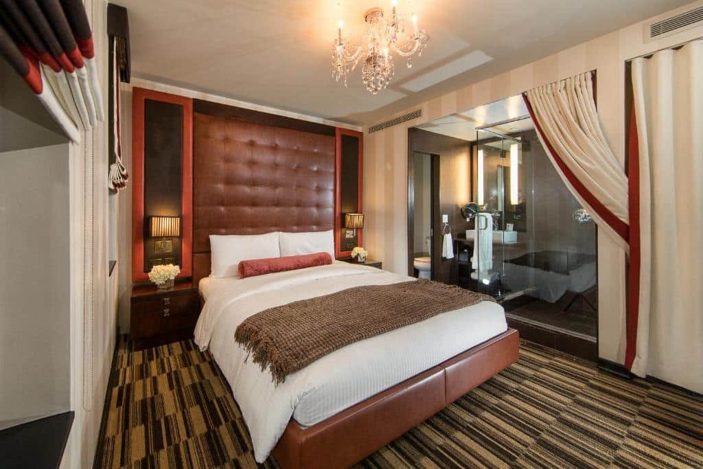 quarto do Sanctuary Hotel New York com uma cama de casal bem no centro da suíte, embaixo de um lustre de cristais pendurado no teto. No lado direito da cama é possível ver uma porta aberta para o vaso sanitário, ao lado da pia com espelho e um box de vidro transparente.